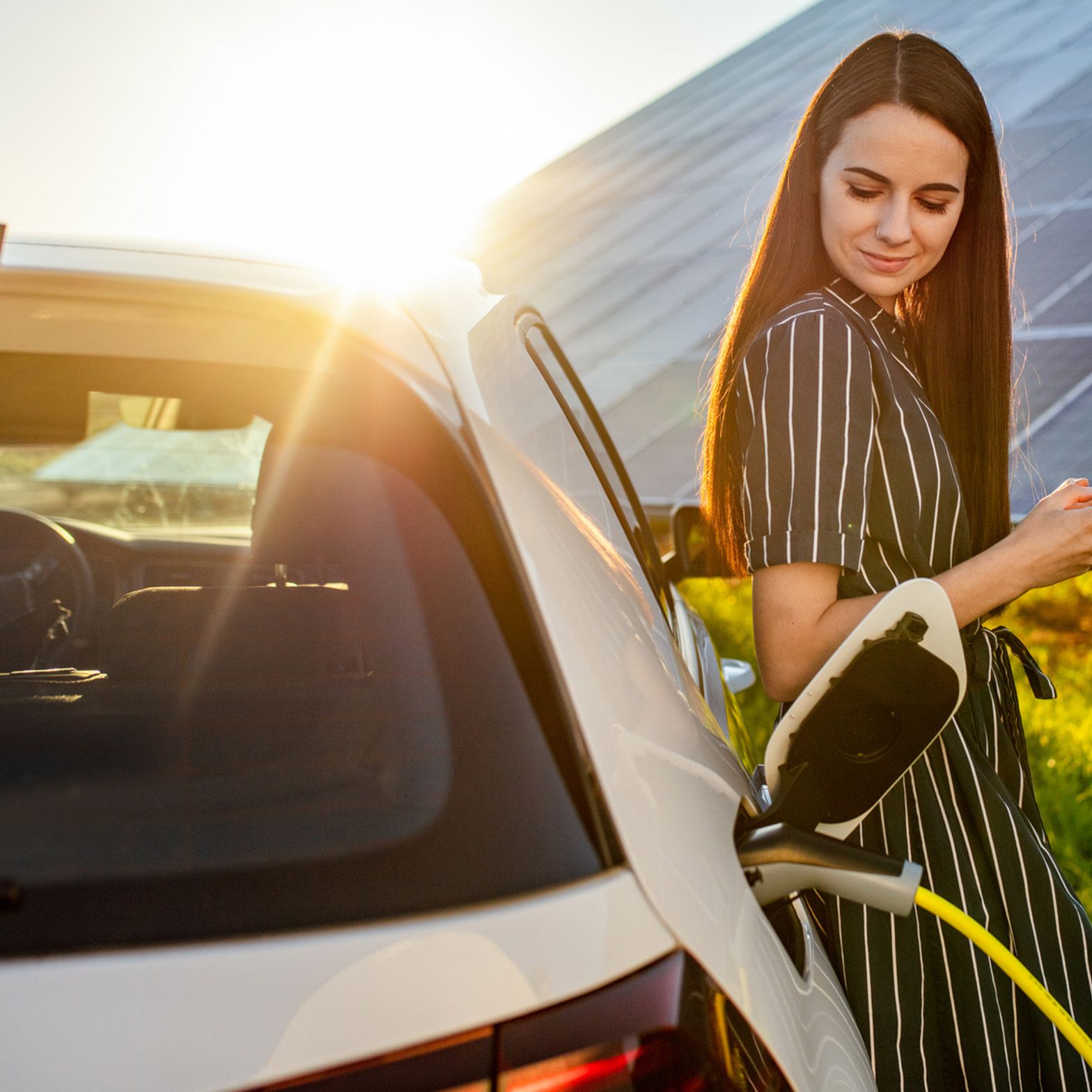 Žena opřená o nabíjející se auto před fotovoltaickým systémem
